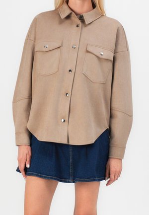 Frau trägt eine beige Jacke mit Knöpfen und Klappentaschen sowie einen dunkelblauen Jeansrock, steht vor einem einfarbigen Hintergrund.