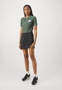 Zelený rebrovaný polo tričko s bielym golierom, skombinované s čiernou sukňou. Model nosí čierne tenisky s bielym piktogramom a ponožky.