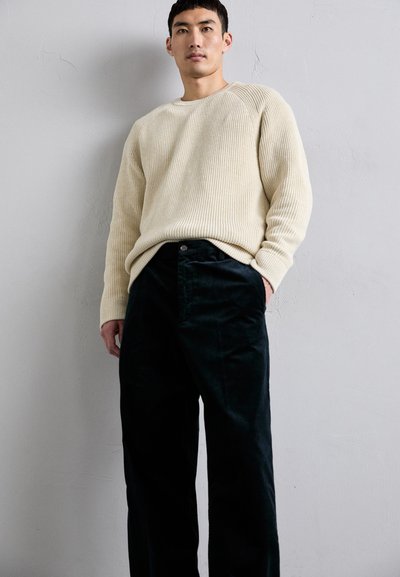 Suéter de punto acanalado en crema combinado con pantalones de pana negros. El suéter tiene un escote redondo y un corte relajado, mientras que los pantalones tienen un cierre de botón.
