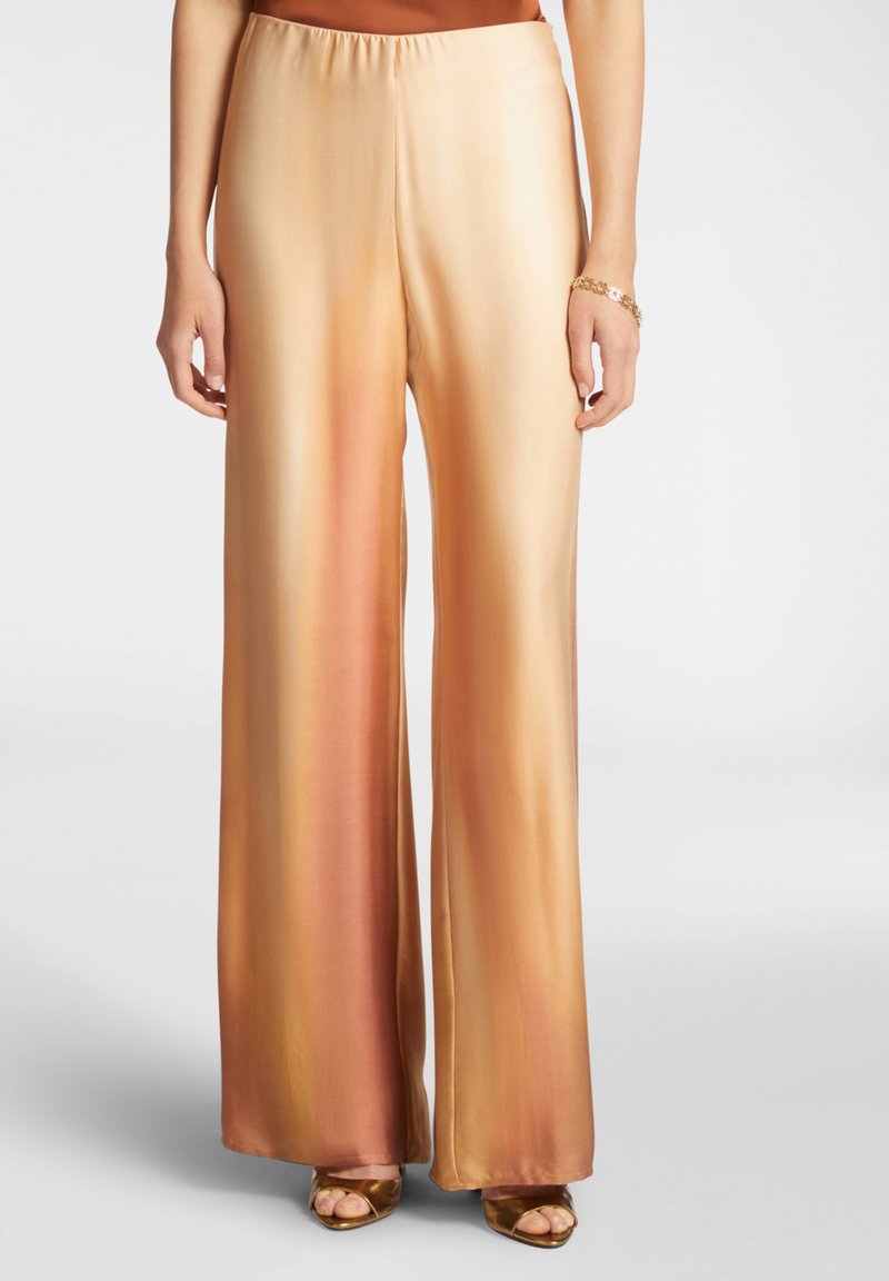 Pantaloni a gamba larga in raso con sfumature di arancione e oro, caratterizzati da una vita elasticizzata e una texture liscia, abbinati a sandali open-toe.