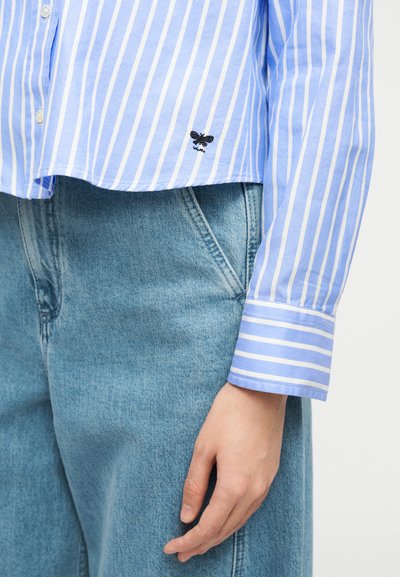 Άτομο που φοράει ανοιχτό μπλε τζιν και πουκάμισο με κάθετες ρίγες σε μπλε και άσπρο, με μικρή μαύρη κεντημένη μέλισσα.