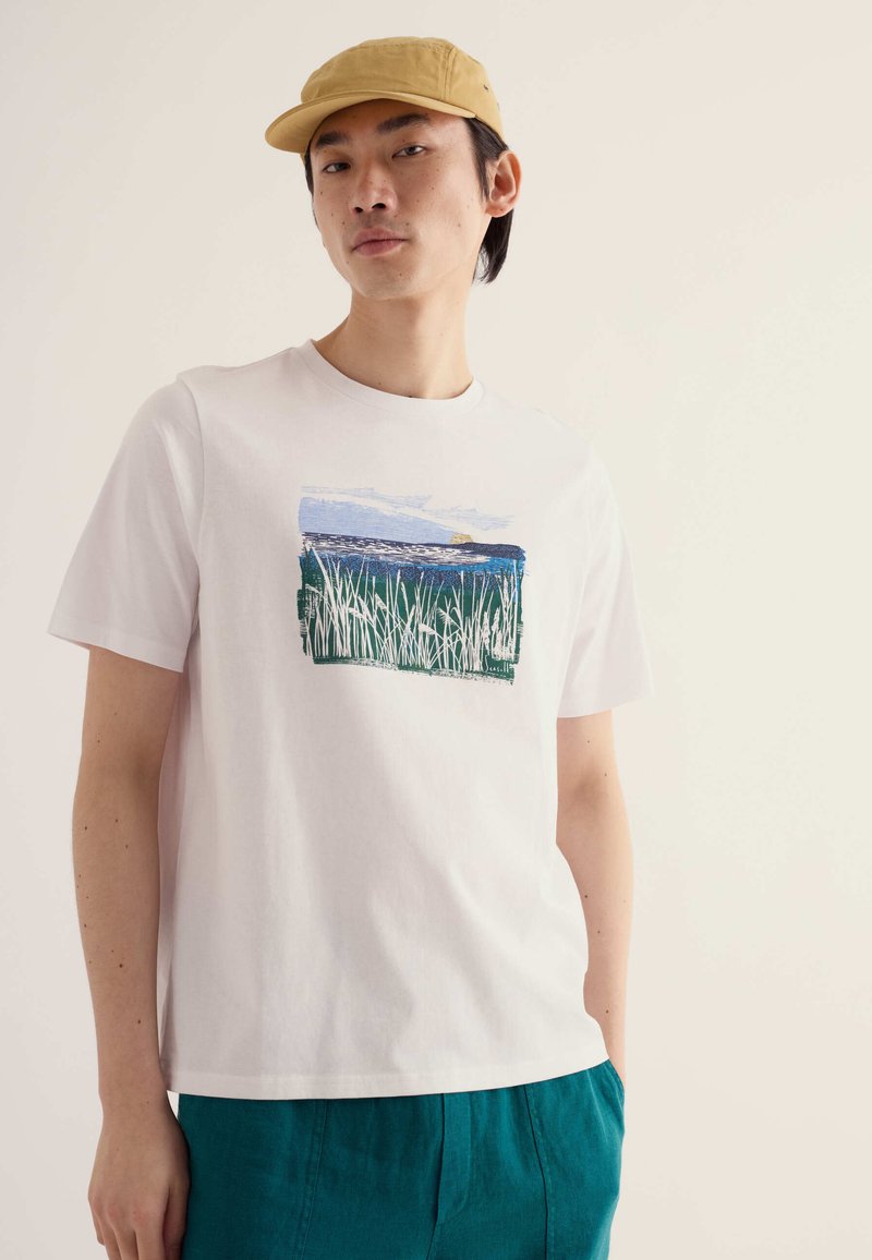 Jeune homme portant une casquette beige, un t-shirt blanc avec un imprimé nature bleu et vert, et un pantalon sarcelle, debout devant un fond clair uni.