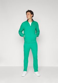 Green tracksuit made of soft fabric. Zip-up jacket with side pockets and contrast piping. Matching pants with tapered legs, both featuring logo accents.