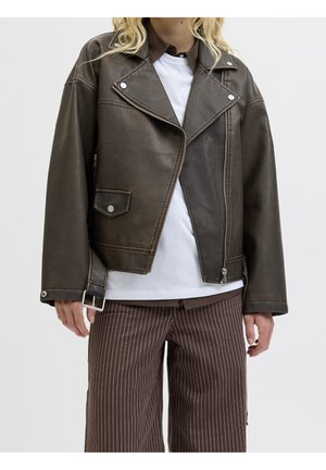 Personne portant une veste de motard en cuir brun foncé sur une chemise blanche et un pantalon marron à larges jambes à fines rayures sur un fond uni.