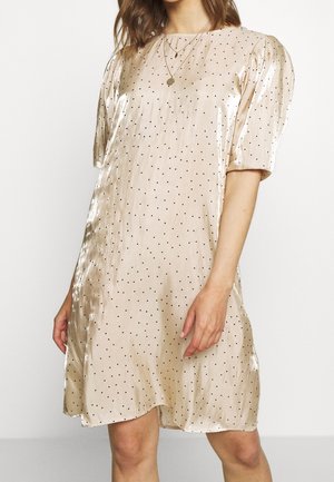 Kvinde iført en skinnende beige kjole med sorte polkaprikker, pufærmer og rund hals, accessoriseret med en rund vedhængs-halskæde.