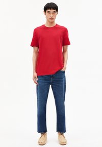 Mann steht mit der Hand in der Tasche, trägt ein rotes T-Shirt mit kurzen Ärmeln, blaue Jeans und beige Mokassins vor einem weißen Hintergrund.