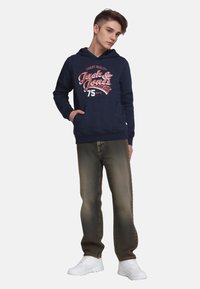 Felpa con cappuccio blu navy con testo grafico e logo sulla parte anteriore, abbinata a jeans di denim marroni sbiaditi e sneakers bianche.