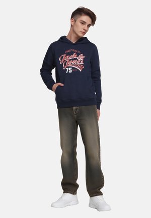 Felpa con cappuccio blu navy con testo grafico e logo sulla parte anteriore, abbinata a jeans di denim marroni sbiaditi e sneakers bianche.