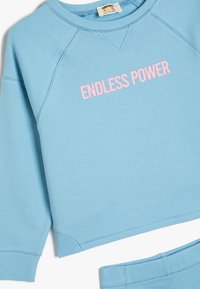 Sudadera de color azul claro con acentos de canalé, que presenta el texto "PODER INFINITO" en rosa. Tejido suave con un ajuste relajado y cuello redondo.