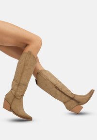 Botas de vaquero de ante en tono beige con punta afilada y tacón de madera en bloque. Presentan costuras decorativas y un cierre lateral con cremallera para facilitar su uso.