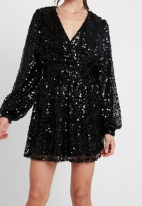 Kvinna i en svart paljetterad långärmad mini-klänning med djup V-ringning och insvängd midja mot en enfärgad bakgrund.