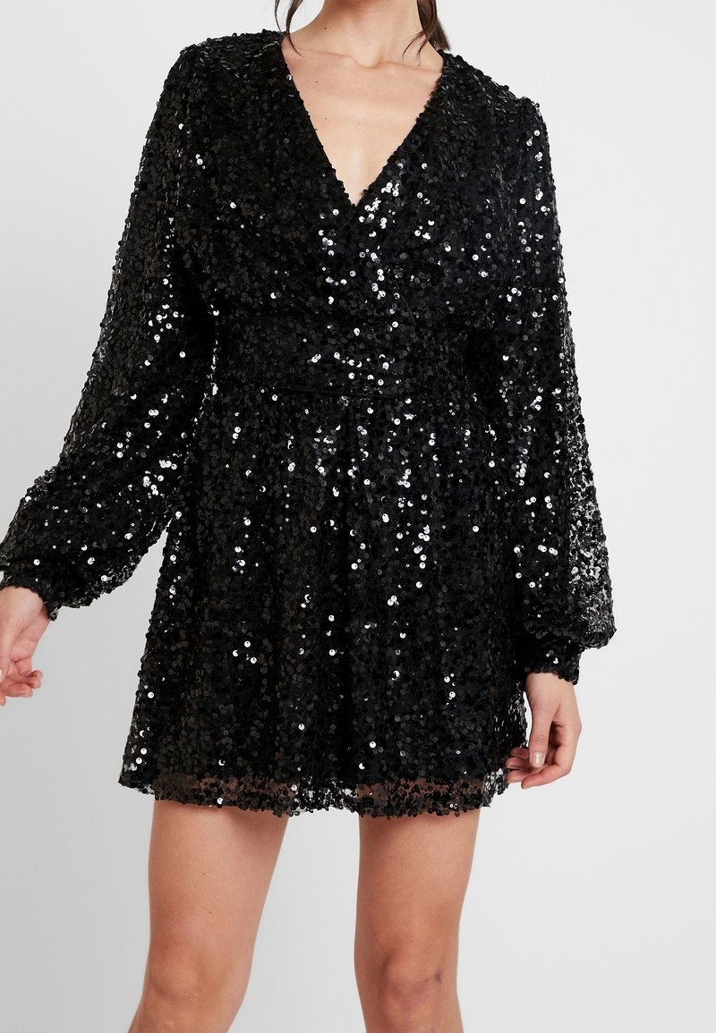 Kvinna i en svart paljetterad långärmad mini-klänning med djup V-ringning och insvängd midja mot en enfärgad bakgrund.