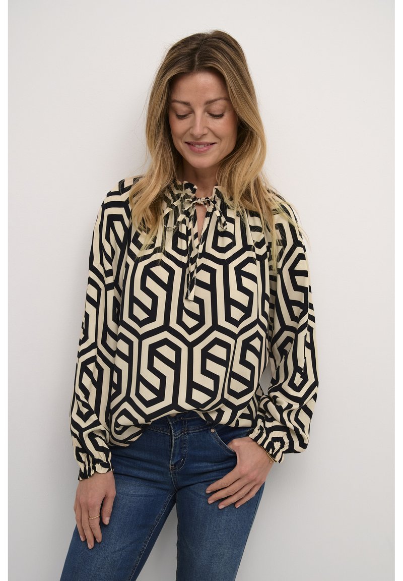 Blusa a maniche lunghe color crema con un motivo geometrico nero. Presenta un colletto legato e polsini elasticizzati. Abbinata a denim blu.