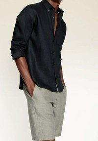 Chemise noire en lin avec boutons et manches retroussables, associée à un short en lin gris clair. La tenue présente une coupe décontractée et un design simple.