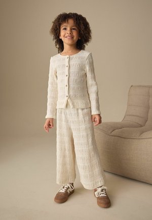 Young child with curly hair wearing textured cream buttoned cardigan and matching wide-leg pants, standing near beige cushioned chair.