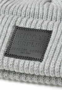 Beanie grigio intrecciato con una trama a coste, caratterizzato da una patch in pelle nera con la scritta "Minoti Surplus Apparel Goods" e cuciture decorative.