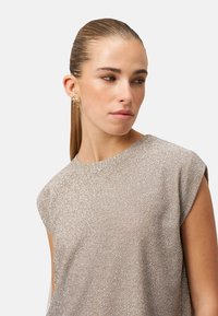 Kobieta z zaczesanymi do tyłu włosami, ubrana w błyszczącą, bezrękawnikową bluzkę oraz złote ozdobne kolczyki, patrząca w lewo na tle gładkiego tła.