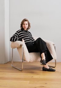 Haut à rayures noir et blanc avec des boutons, pantalon large noir et mocassins noirs. Assis sur une chaise en peluche de couleur crème avec un cadre en chrome.