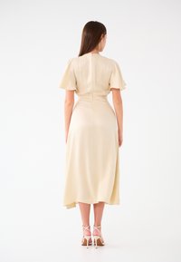 Midikleid in hellbeige, mit kurzen Rüschensäumen, tailliertem Schnitt und einem schwingenden Rock mit asymmetrischem Saum. Rückenzipper-Detail.