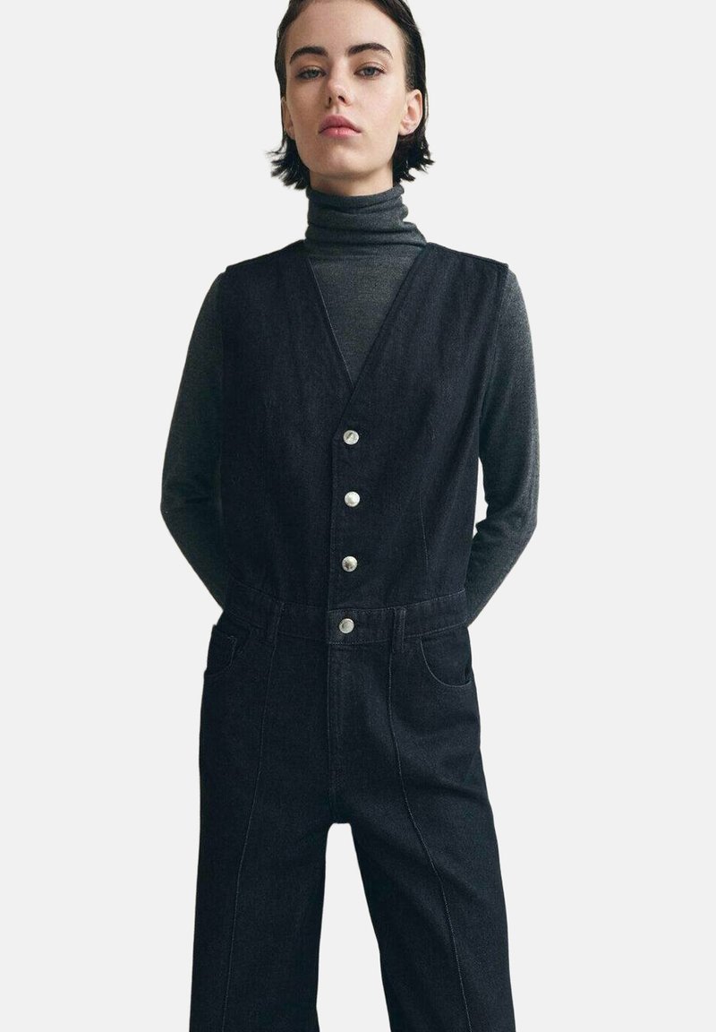 Vestido de peto de mezclilla negra con un escote en V profundo, botones plateados y bolsillos laterales. Diseño minimalista con detalles de costuras en contraste.