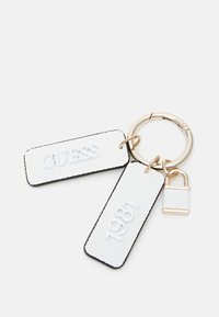 Llavero con dos etiquetas blancas texturizadas, con las palabras en relieve "GUESS" y "1981", sujetas a un anillo metálico dorado y un pequeño candado como adorno.