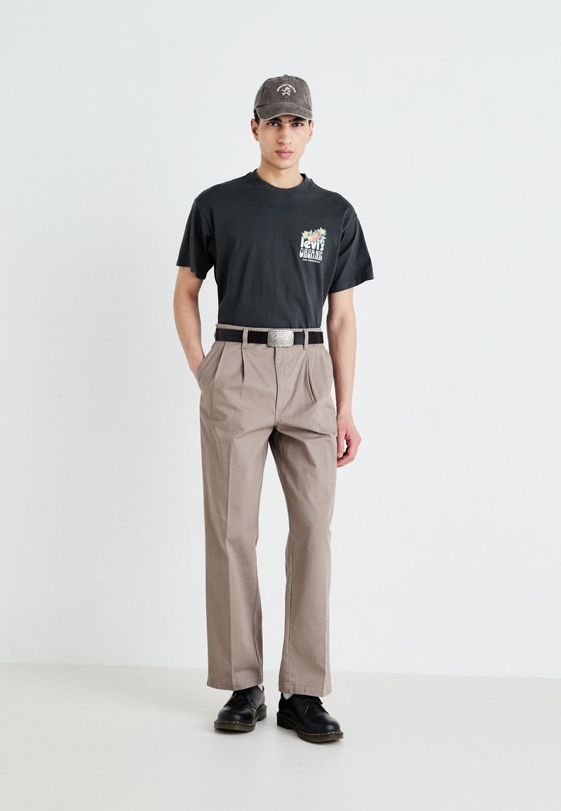 T-shirt graphique noir à manches courtes, pantalon plissé marron clair, ceinture noire avec boucle en argent, et chaussures noires. Le modèle se tient sur un sol clair.