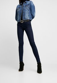 Person som bär mörka skinny jeans, svarta klackstövlar, ett nitarbälte, svart topp och en blå denimjacka mot en enkel bakgrund.