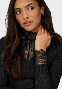Maglietta nera a maniche lunghe con tessuto a costine e delicati accenti in pizzo nero sul collo e sui polsini. La modella ha i capelli lunghi e scuri e un trucco neutro.
