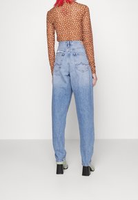 Top marrón de manga larga con estampado floral, combinado con vaqueros holgados de talle alto en azul claro. Zapatos de tacón verdes, con bolsillos traseros visibles.