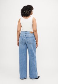 Zili zili džinsi ar vidēju jostasvietu, ar brūnu plāksteri aizmugurējā jostas vietā un redzamām šuvju detaļām. Redzama balta topiņa.