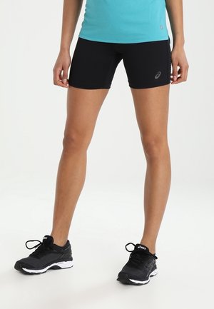 Person trägt schwarze Sportshorts und schwarze Laufschuhe, steht mit leicht gespreizten Beinen vor einem schlichten weißen Hintergrund.