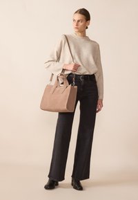 Borsa in suede tan con manici doppi e tracolla regolabile, abbinata a un maglione chiaro e jeans svasati scuri, su uno sfondo neutro.
