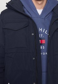 Marinblå pufferjacka med dragkedja fram, två bröstfickor och huva. Bärs över en blå hoodie med en logotyp. Mjuk, texturerad tyg.