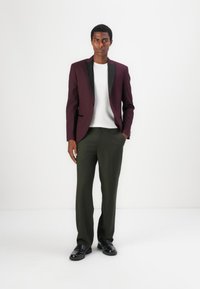 Bordeauxfarbener Blazer mit schwarzen Revers, weißem Hemd, olivgrüner Hose und schwarzen Schuhen. Slim Fit, glattes Material und maßgeschneiderte Optik.