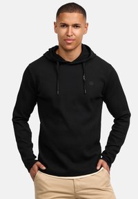 Sudadera negra con costillas y capucha ajustable, con mangas largas y un logo en el pecho. Combinada con pantalones beige claros.