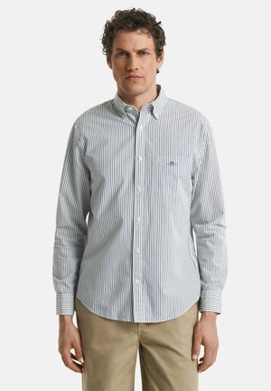Chemise à rayures bleu et blanc avec des poignets ajustables, une poche avant, et un col classique, portée avec un pantalon beige.