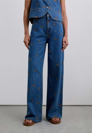 Jeans de pierna ancha azules con bordados florales marrones, combinados con un chaleco bordado a juego y sandalias marrones sobre un suelo de hormigón.