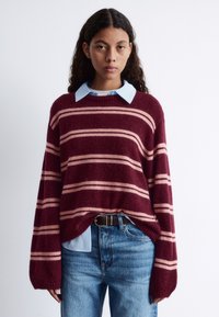 Bordeaux gestreifter Pullover mit rosa Streifen, grob gestrickt, lockere Passform. Getragen über einem hellblauen Hemd mit Kragen, kombiniert mit blauen Jeans und einem schwarzen Gürtel.