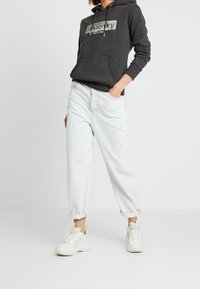 Kohlefarbener Hoodie mit Logo auf der Vorderseite, kombiniert mit hellblauen, hochgekrempelten Jeans. Das Modell trägt weiße Sneaker vor einfachem weißem Hintergrund.