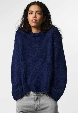 Jeune femme aux cheveux bouclés et foncés portant un pull ample et épais bleu marine et un jean gris clair sur un fond uni.