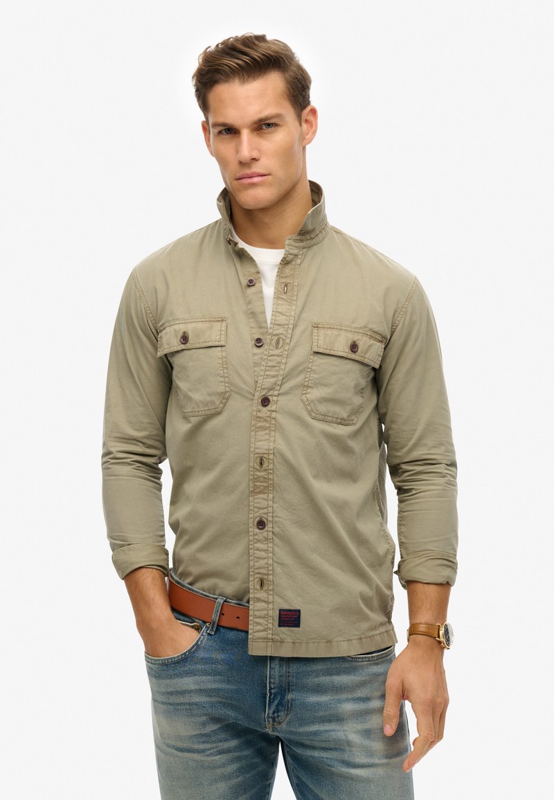 Khaki button-up shirt met twee borstzakken, lange mouwen en een staande kraag. Draagt met blauwe spijkerbroek en een leren riem.