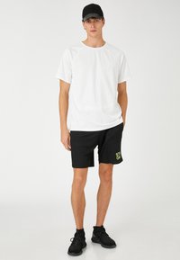 Camiseta blanca de manga corta combinada con pantalones cortos negros que presentan un gráfico de neón verde que dice "STRONG". El modelo lleva una gorra negra y zapatillas deportivas.