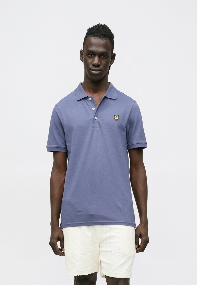 Soe sinine polo särk kraega, ees kolm nuppu ja väike kollane logo vasakul rinnal; kombineeritud valgete tekstuursete lühikestega.