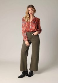 Rode blouse met bloemenpatroon en een V-hals, gecombineerd met olijfgroene wijde broekspijpen. Model draagt zwarte enkellaarsjes.