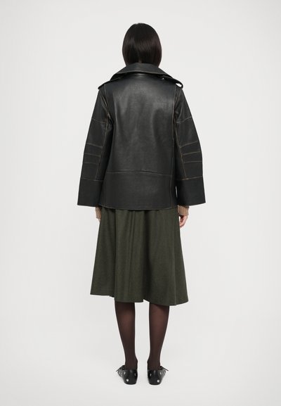 Must Black leather jacket with wide sleeves and contrasting brown stitching, paired with a dark green skirt and black shoes with studs.