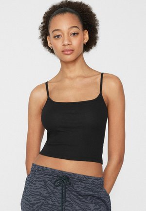 Jeune femme aux cheveux bouclés portant un débardeur noir court et un pantalon gris à motif avec cordon de serrage, debout devant un fond uni.