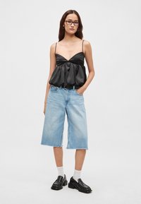 Czarny satynowy top na ramiączkach z marszczoną talią, zestawiony z jasnoniebieskimi szortami jeansowymi do kolan. Modelka ma na sobie czarne buty i białe skarpetki.