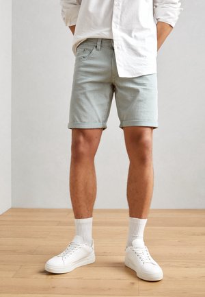 Homme portant un short en denim gris clair, une chemise blanche à manches longues, des chaussettes blanches et des baskets blanches, debout sur un sol en bois.