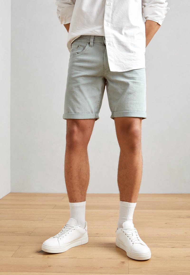 Homme portant un short en denim gris clair, une chemise blanche à manches longues, des chaussettes blanches et des baskets blanches, debout sur un sol en bois.