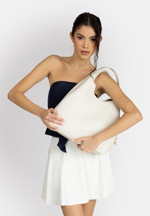 Giovane donna con capelli scuri tiene una grande borsa bianca, indossando un top blu navy senza spalline e una gonna plissettata bianca su sfondo semplice.